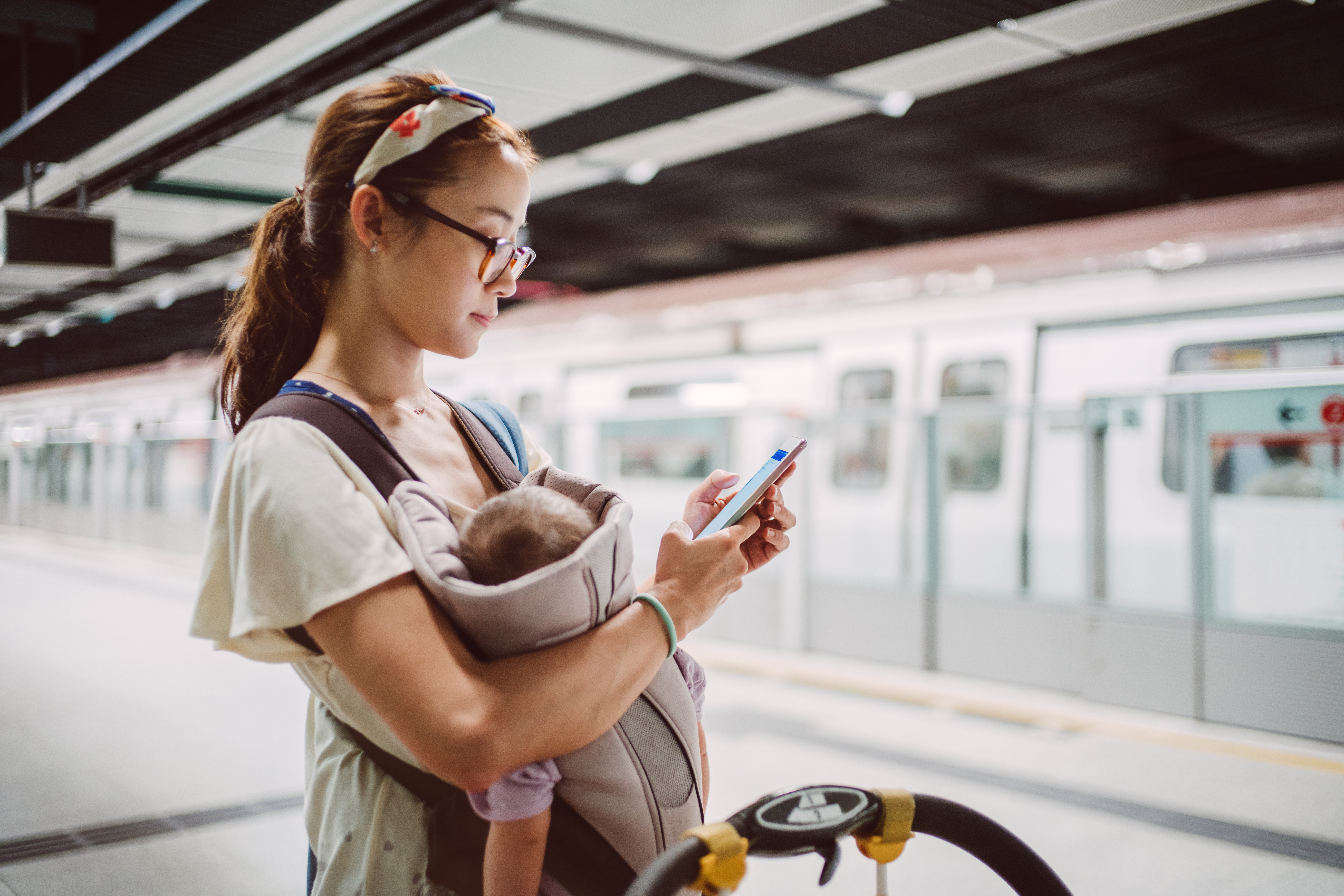 Pretty young mom carrying baby in a baby carrier using smartphone next to a baby stroller on the train platform.